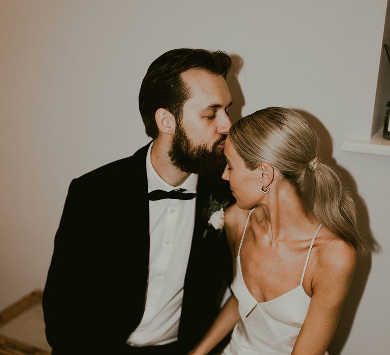 Groom wearing a black tuxedo with white rose buttonhole kisses th bride on the forehead who wears a fitted white gown and small gold hoops 