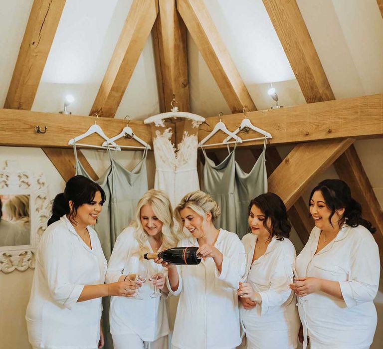 Bride and bridesmaids in matching white pyjama sets and sage green and white fluffy slipper sliders having Champagne before wedding 