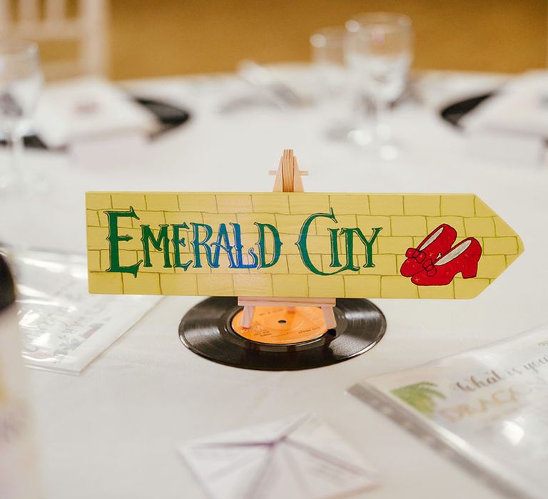 Film-inspired wedding table number in the style of The Wizard Of Oz; a yellow brick patterned arrow sign with ruby red slippers and "Emerald City" in green lettering 