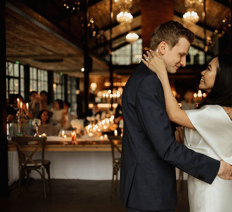 The bride in a sati finish Eva Lendel wedding dress leaning up to embrace the groom 