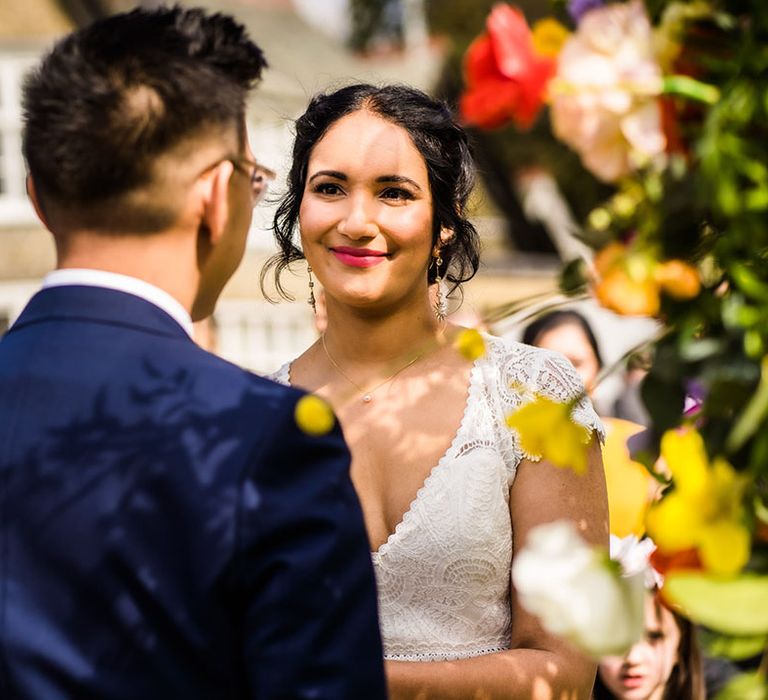 Bride in lace wedding dress looks lovingly toward her groom during outdoor wedding ceremony