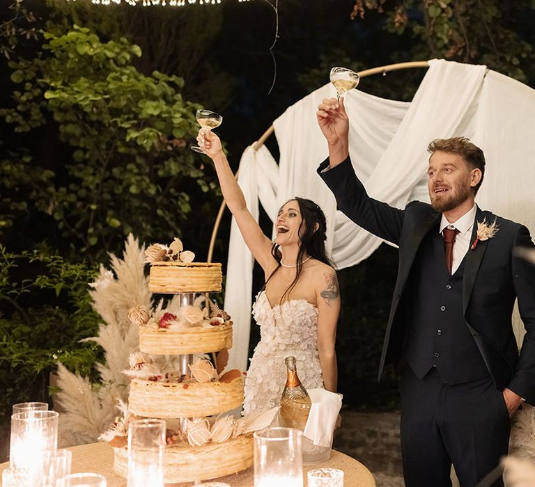 Bride & groom cheers in front of archway complete with white drapes beside Opera wedding cake