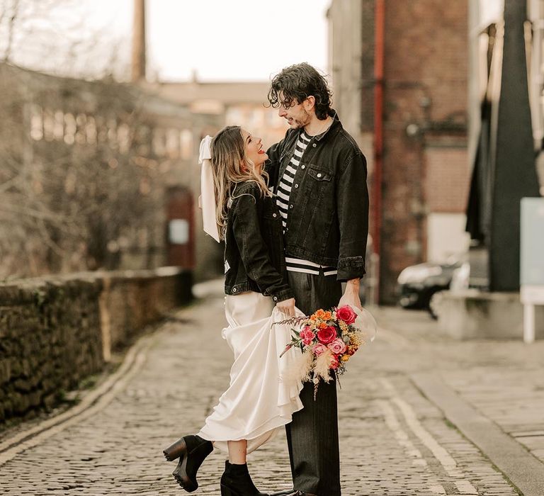 Bride in personalised black denim jacket and satin wedding dress looking up at groom in black and white striped shirt, grey pinstripe trousers, Dr Martens boots and black personalised denim jacket outside The Chimney House wedding venue 