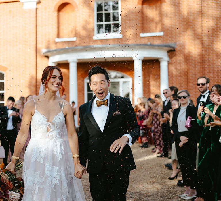 Bride in Madi Lane Bridal wedding dress and her groom in black tie walk through confetti exit 