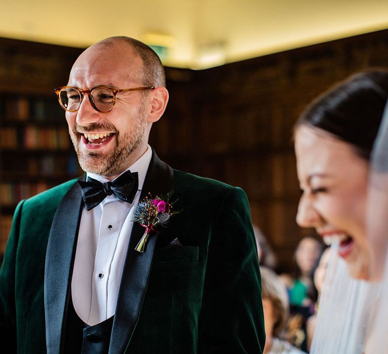 Groom in a velvet green tuxedo with tortoise shell pattern glasses laughing with the bride laughing during their ceremony 