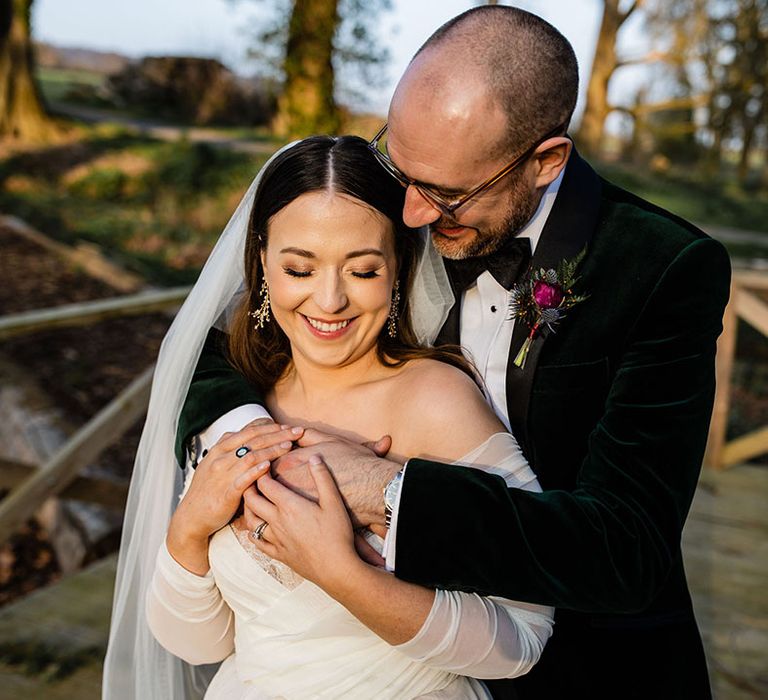 Groom in green velvet suit jacket embracing the bride from behind wearing an off the shoulder long sleeve wedding dress 