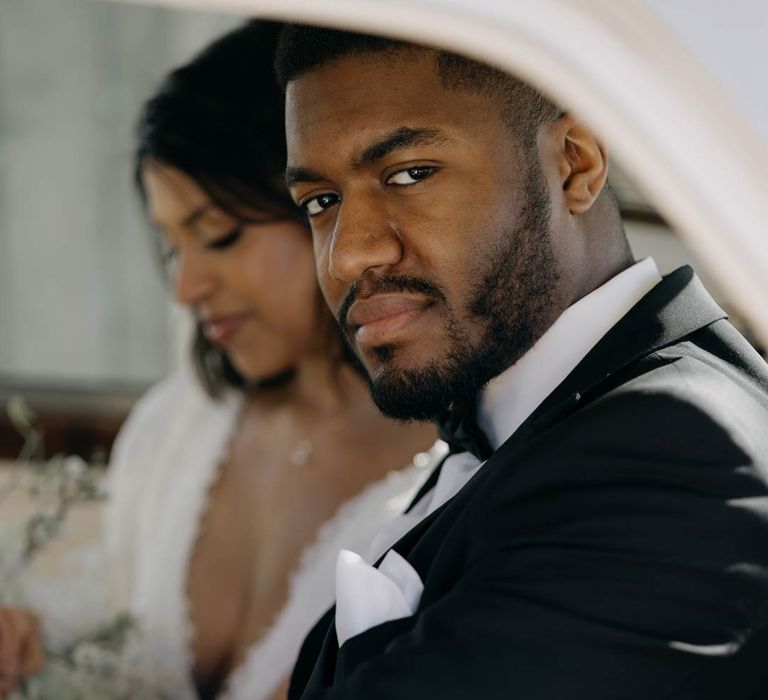 Groom wearing classic black tuxedo, black bowtie and white pocket square sitting in a classic white wedding car with bride