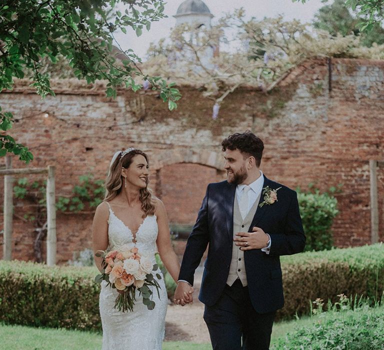 The groom in a dark blue suit jacket and pale waistcoat and tie walks along with the bride in a fitted lace wedding dress with a tiara and veil 