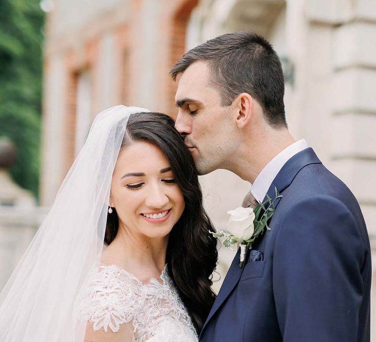 Groom in a navy suit kissing his bride's head in a fitted wedding dress with lace illusion neckline 