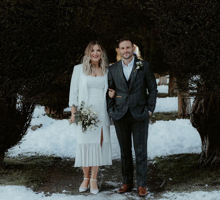 Bride with bright red lipstick on for winter Christmas wedding in ruffle wedding dress and blazer poses with the groom in a dark grey suit for Stroud Registry Office 