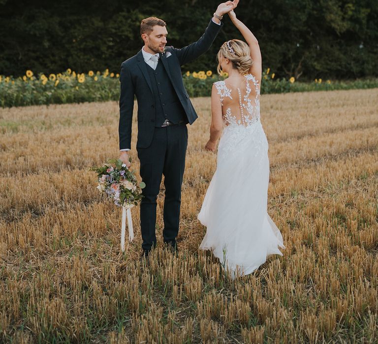 The groom in a three piece navy wedding suit spins the bride around in the fields surrounding Dove Barn Weddings 