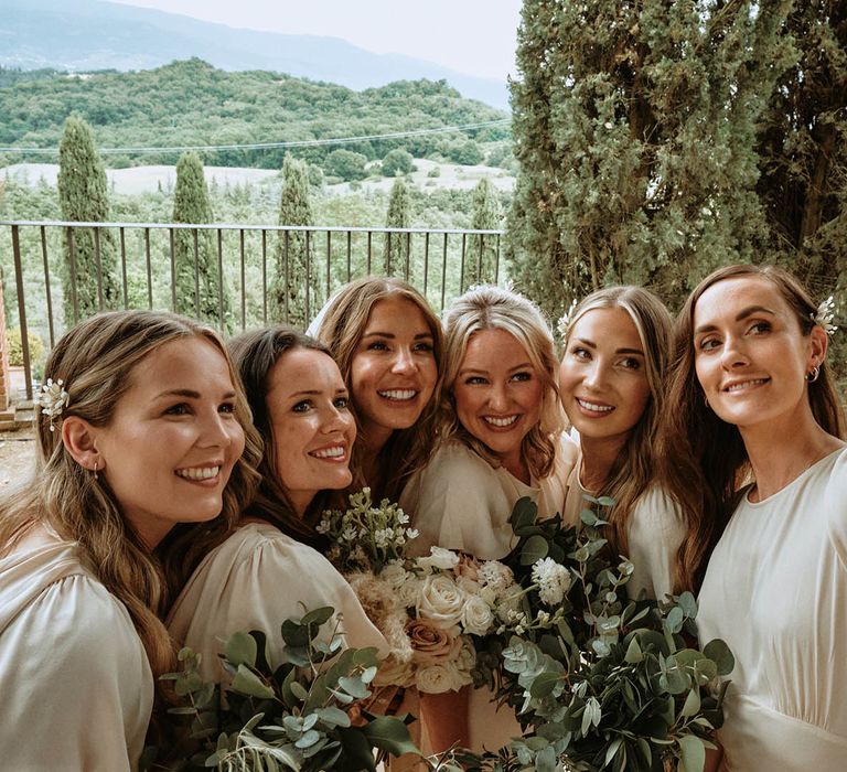 Bride and bridesmaids in off-white satin bridesmaids dresses taking a selfie while holding eucalyptus bridesmaid bouquets