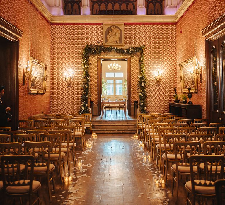 Aisle of Grittleton House wedding venue with high columns and ceilings, wooden chairs, warm cosy lighting and slim flower arch going around the door 