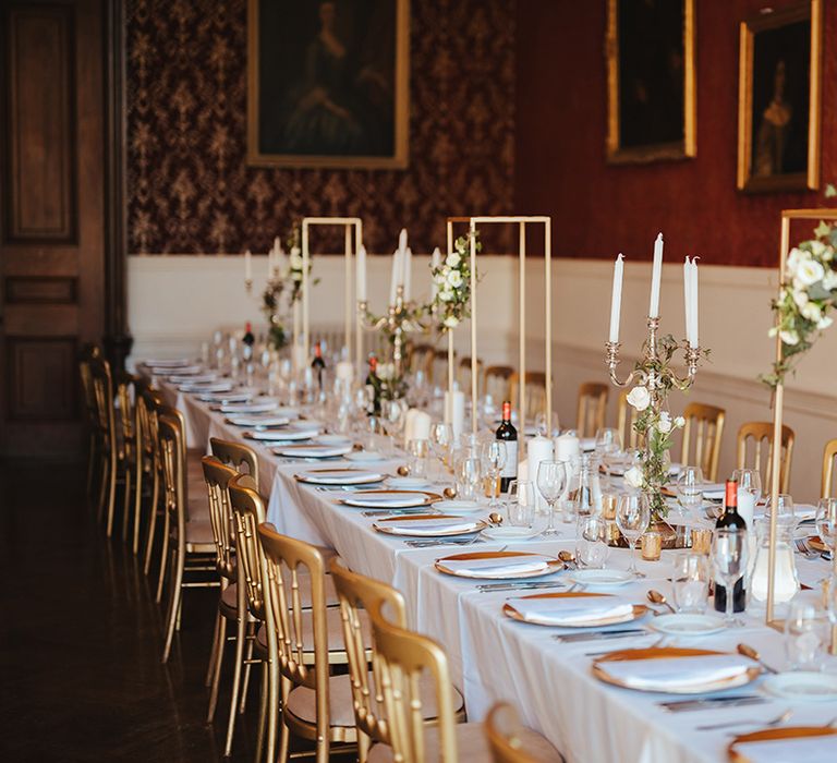 Classic wedding tablescape with white table cloth, tapered candles in gold candelabras, gold frame decorations, gold plates, gold cutlery and dried flower and white rose floral arrangements at Grittleton House 