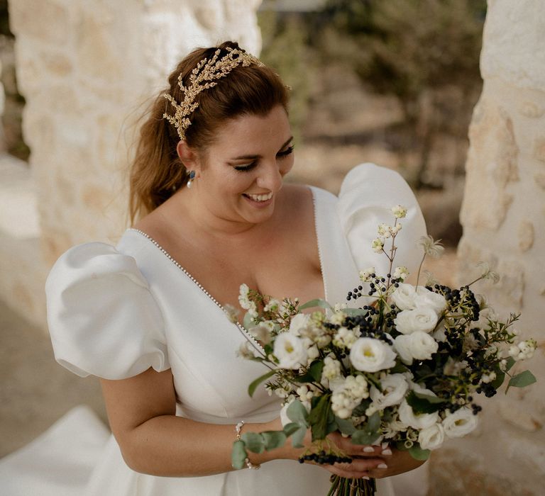 Bride carrying bridal bouqet that features wihte berries, italian ranunculus and eucalyptus whilst wearing a stain puff-sleeved wedding dress, peal earrings and gold leaf bridal grown