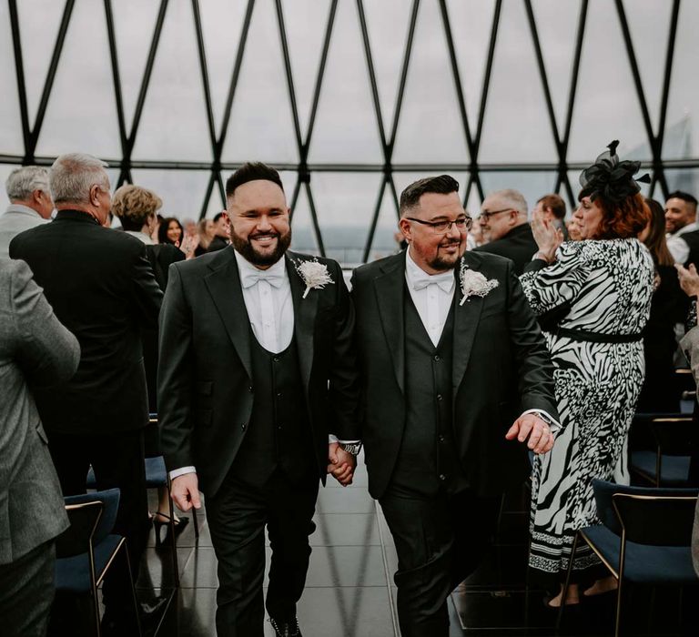 Grooms in three piece black tuxedos with off white bow ties and white garden rose, lavender twig and dried foliage boutonnieres walking back down the aisle at The Gherkin wedding venue London