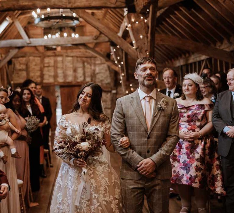 Bride in long sleeve lace illusion sleeve wedding dress with overlay at the end of the alter of Bassmead Manor Barns seeing bride in maroon bridal suit with grey waistcoat, dusky pink tie and autumnal dried flower boutonniere 