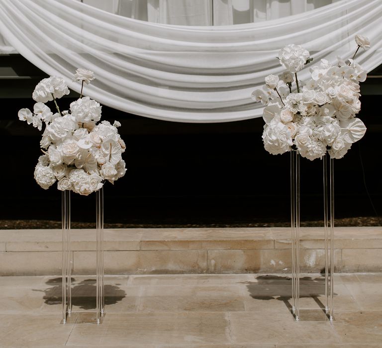 White wedding flower arrangements with orchids, roses, and anthuriums on clear acrylic podiums with white drapery decorating the venue 