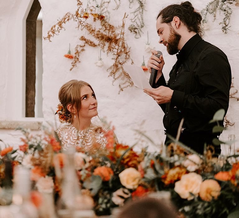 The groom in his all black outfit stands up to read out his wedding speech with the bride listening intently 