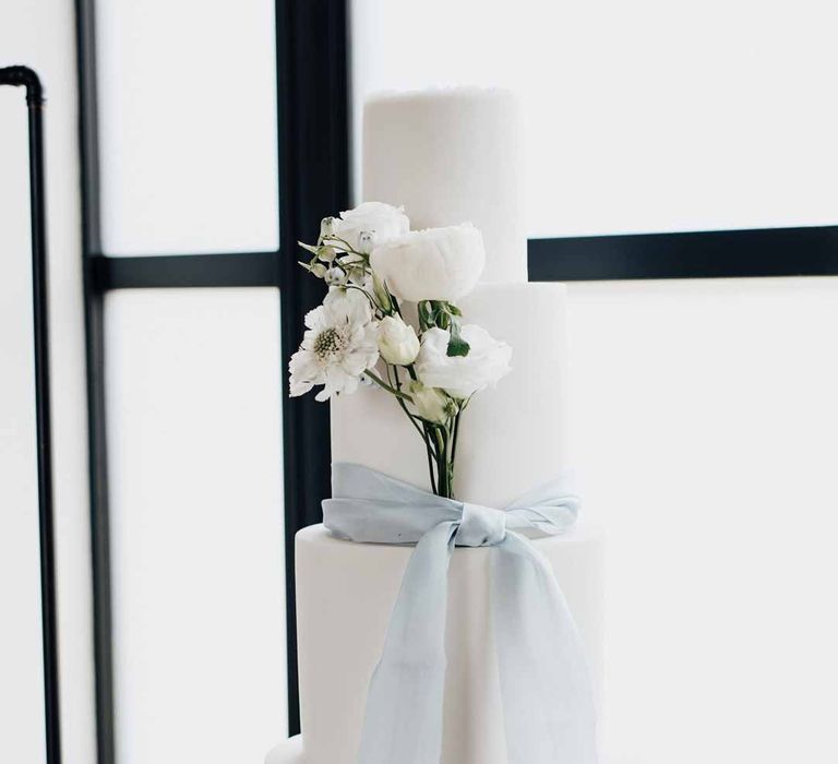 Three tier iced wedding cake in white with baby blue ribbon and white flower cake decorations on clear cake stand 