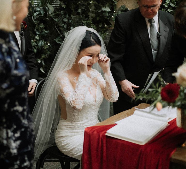 The bride in a long sleeve Pronovias wedding dress and white veil wipes her eyes as she gets emotional signing the wedding register 