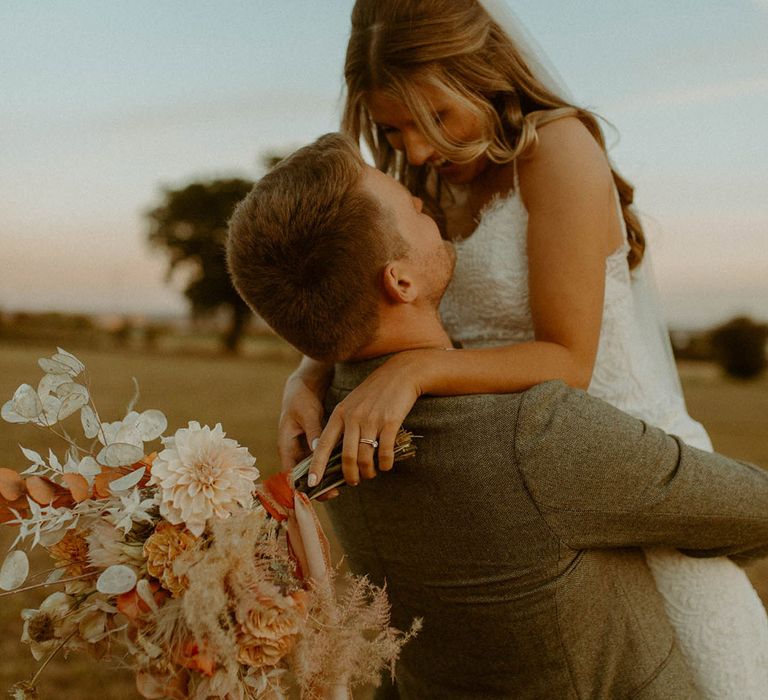 Bride with hair in a half up half down hairstyle being lifted for intimate embrace by the groom in a grey wedding suit with an orange and white bouquet 