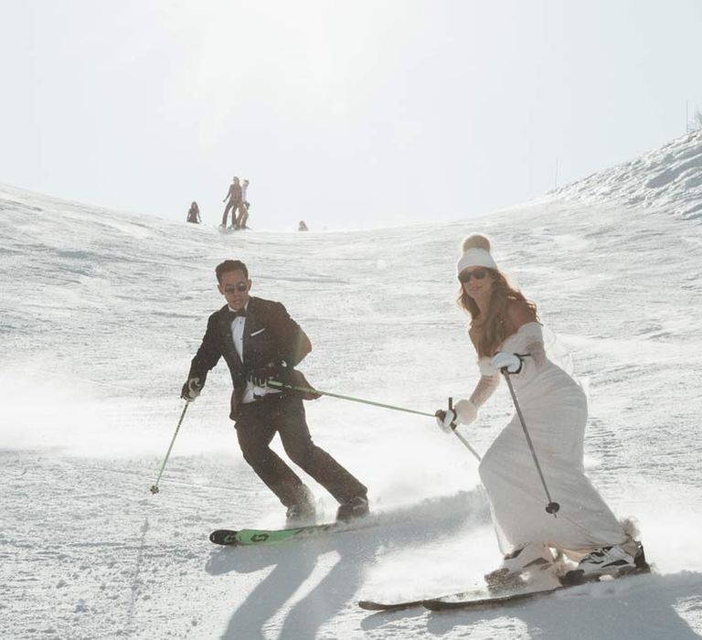 Bride in off shoulder sparkly mermaid wedding dress, wooly bobble hat and sunglasses skiing with groom in classic black tuxedo, black bowtie, sunglasses and white garden rose boutonniere at French ski wedding