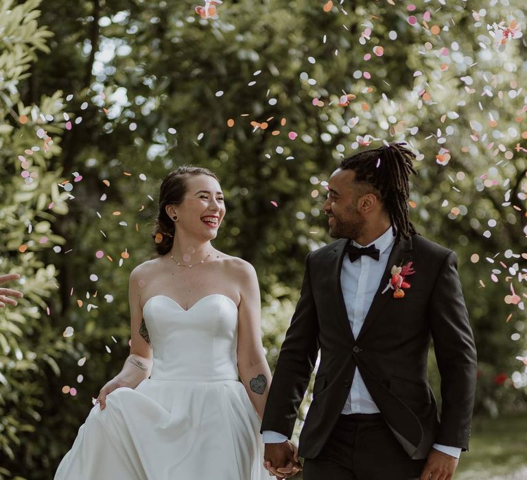 Groom in classic black tuxedo with bowtie and pink and orange wedding boutonniere walking with bride in strapless sweetheart neckline wedding dress walking through colourful wedding confetti