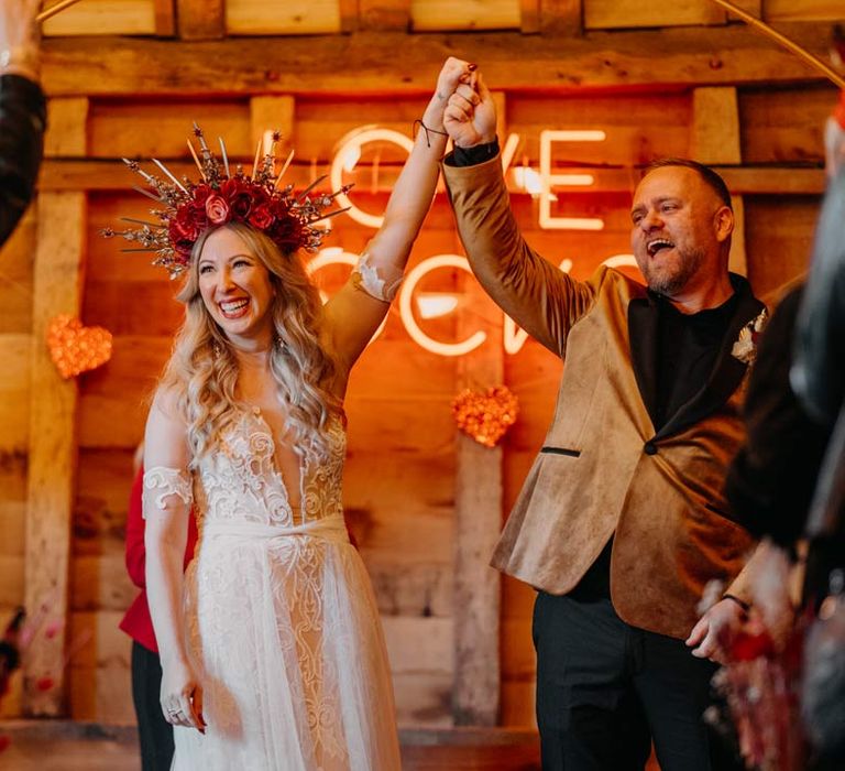 Groom in crushed velvet burnt orange grooms blazer and dried flower boutonniere holding bride's hand up wearing off shoulder lace wedding dress with corset style top and red rose and gold halo bridal crown at Southlands Barn