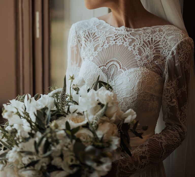 bride in a long sleeve lace wedding dress looking out the window holding a white and green wedding bouquet 