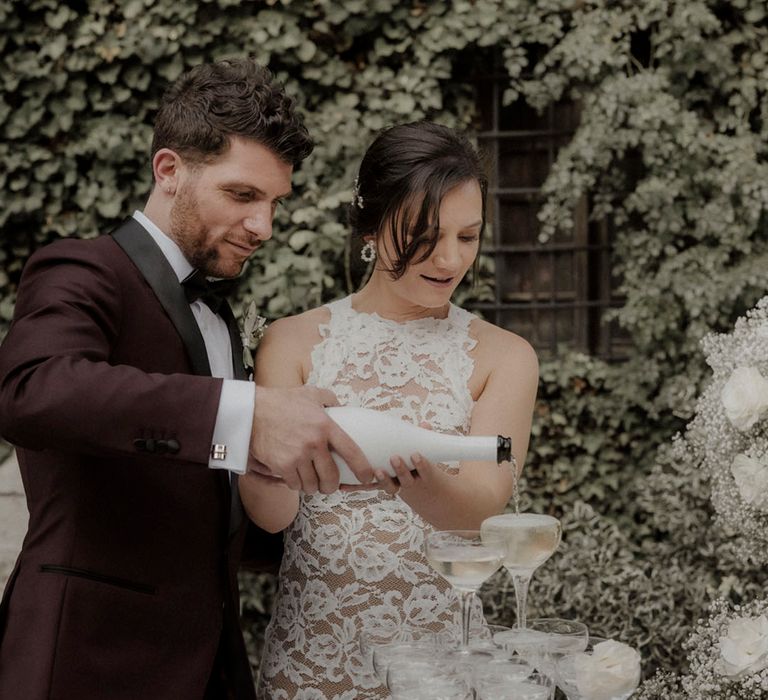 Groom in burgundy suit with bride in long sleeve lace wedding dress pouring champagne onto champagne tower 