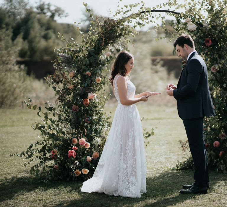 Bride and groom exchanging rings in front of a floral moon gate by Number Twenty Seven