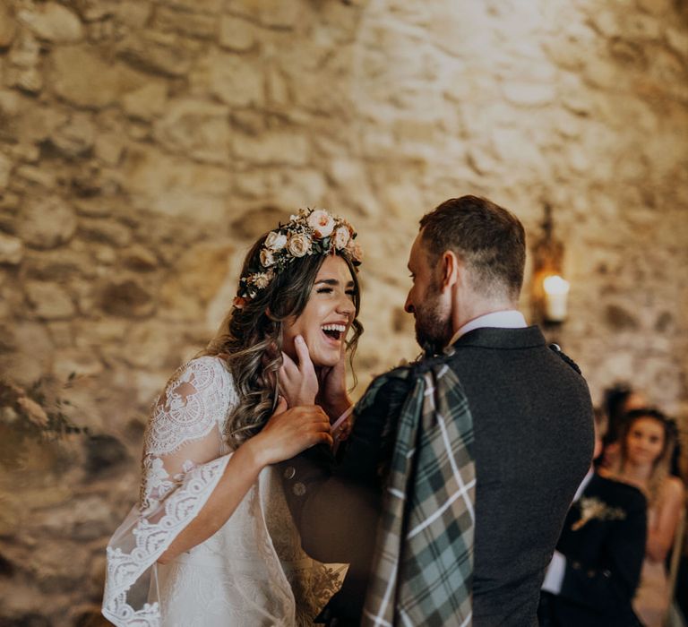 Smiling groom in kilt holds the face of laughing boho bride in pink rose flower crown 