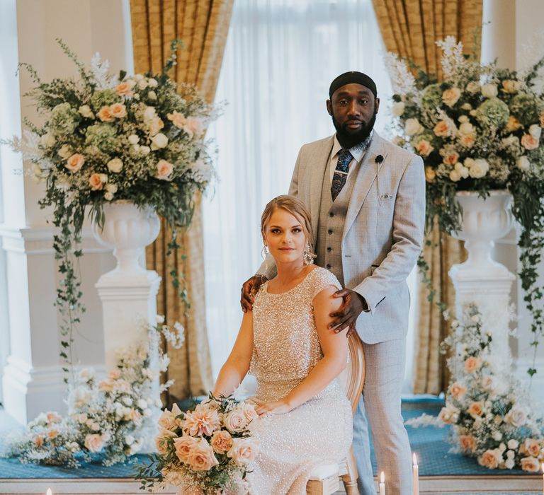 Groom in light grey suit standing next to his bride in sparkly separates surrounded by peach flowers and candles at The Landmark London