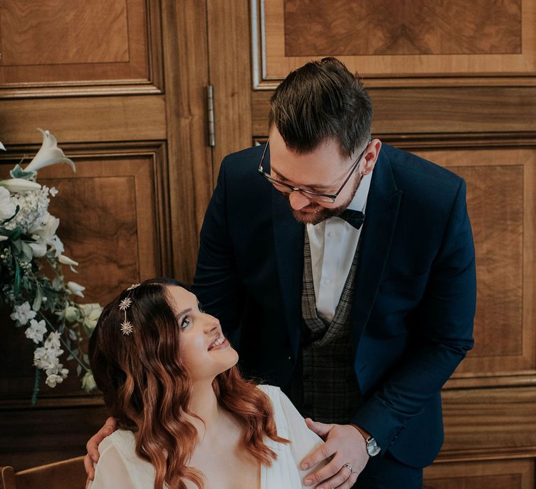 Bride wearing celestial headband looks up at groom in navy suit whilst signing wedding registry 