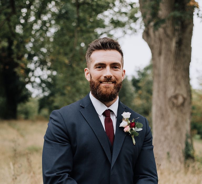 Bearded groom in a navy suit with burgundy tie and white and red buttonhole | Harmonie Photography