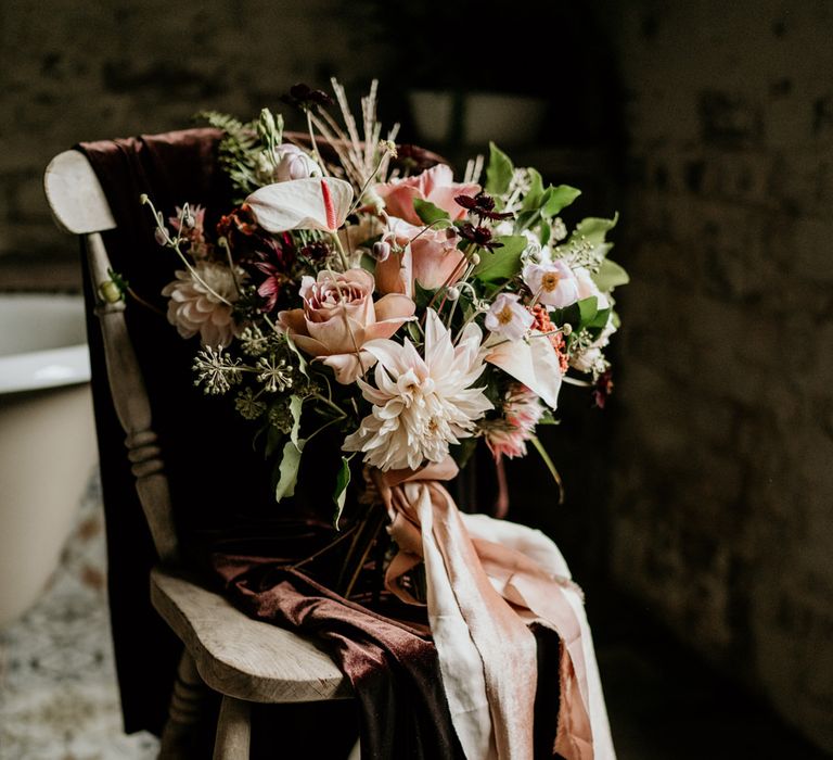 Pink wedding bouquet with dahlias, roses and anthuriums tied with ribbon sitting on a wooden chair 