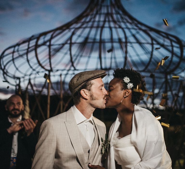 West African bride with short hair kissing her groom in a beige Peaky Blinders wedding suit during the gold confetti moment 