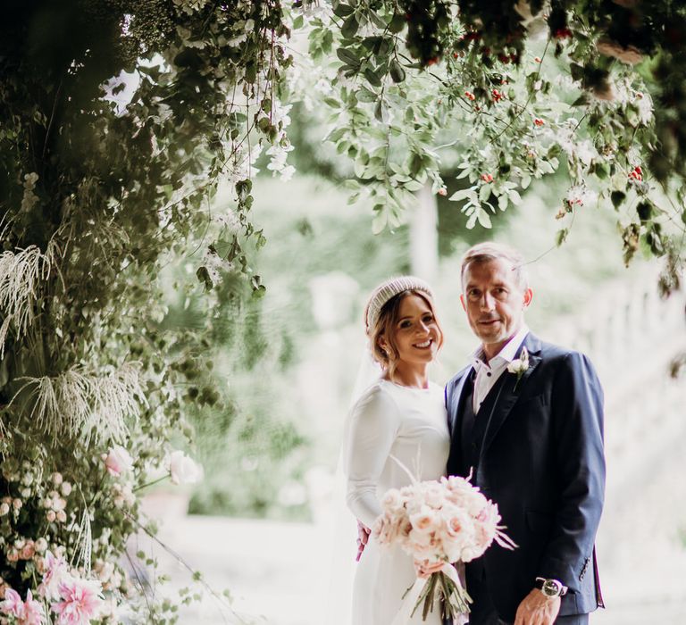 Bride in Elbeth Gillis gown holding blush pink rose wedding bouquet stands outside with groom in navy Cad & the Dandy suit under floral canopy at Euridge Manor wedding