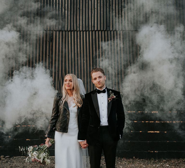 Bride in personalised leather bridal jacket and white Charlie Brear wedding dress holding hands with groom in black Hugo Boss suit with white smoke bomb in background