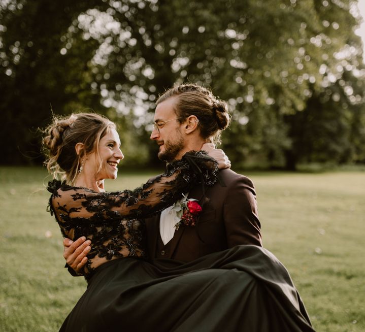 Groom in a burgundy wedding suit holding his bride in a black lace wedding dress for gothic style wedding 