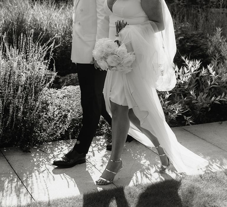 Bride in V neck Made With Love wedding dress and veil holding white rose bouquet walks outside with groom in white double breasted tuxedo jacket