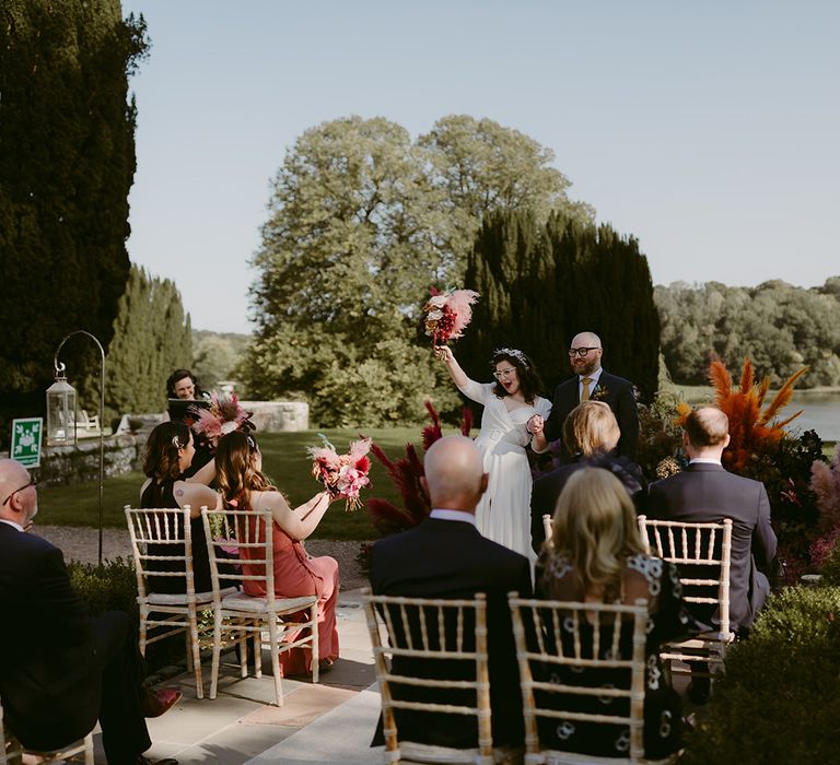 Bride in white Rime Arodaky wedding dress holds up pink pampas grass wedding bouquet in celebration next to groom in navy suit