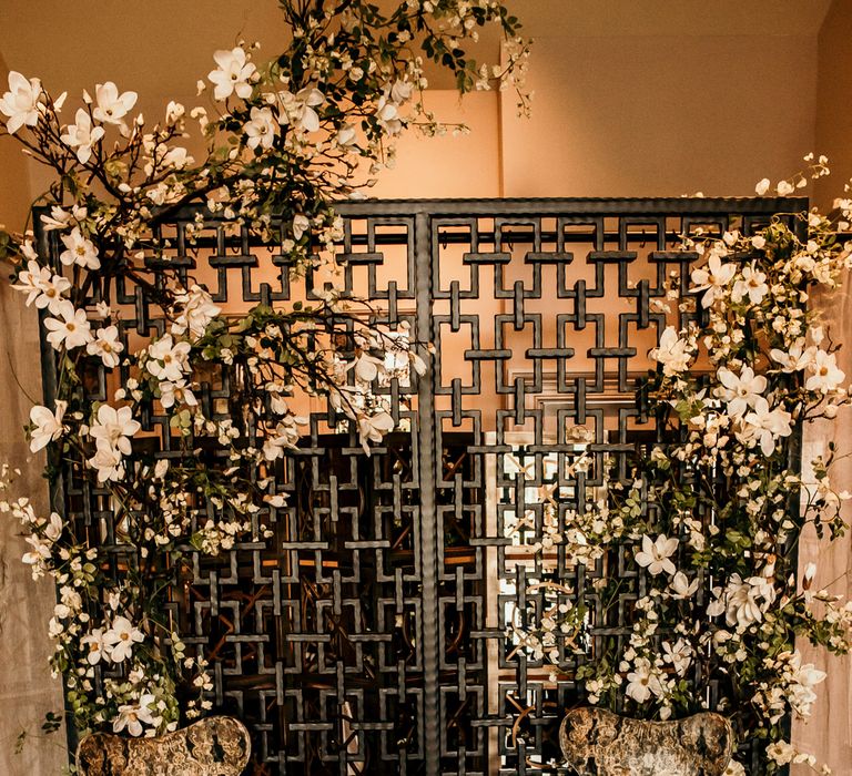 Wooden table with textured chairs sit in front of intricate wall design and floral arch