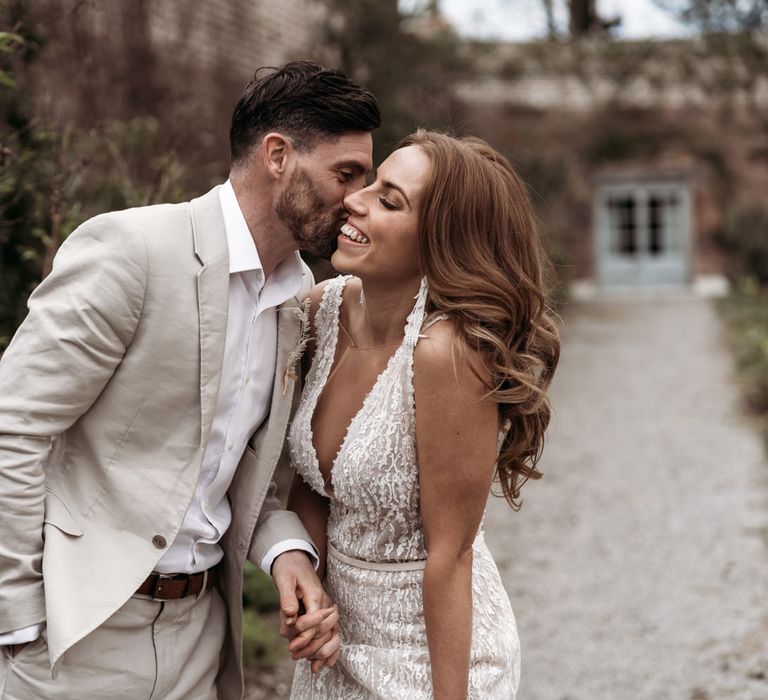 Groom in a beige suit kissing his bride on the cheek with long wavy hair and tassel earrings 