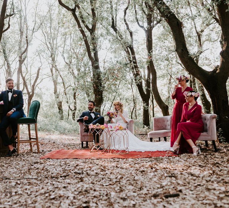 Bride in long sleeved lace boho dress and flower crown sits on pink velvet sofa with groom in navy suit in woodland with wedding guests sat on various velvet seats at enchanted forest wedding in Italy