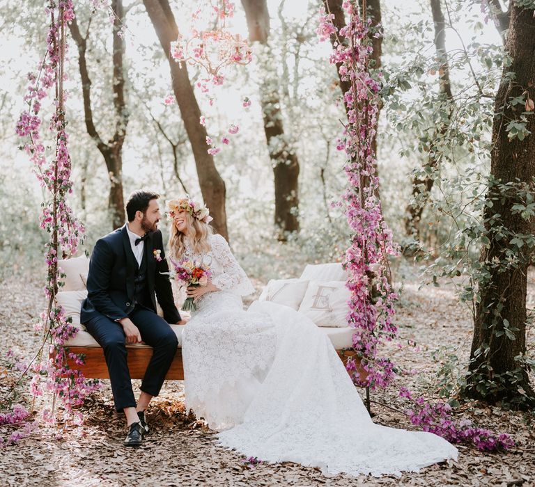 Bride in long lace boho wedding dress and flower crown lounges on outside day bed swing in forest with groom in navy suit and bow tie at enchanted forest wedding in Italy