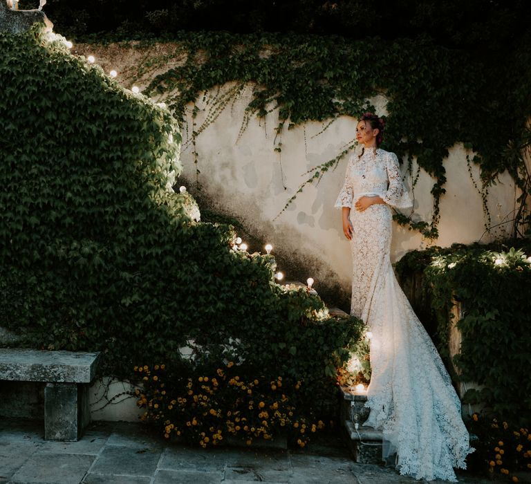 Bride in long sleeved boho lace wedding dress with train stands on fairy-lit steps covered in ivy at enchanted forest wedding in Italy