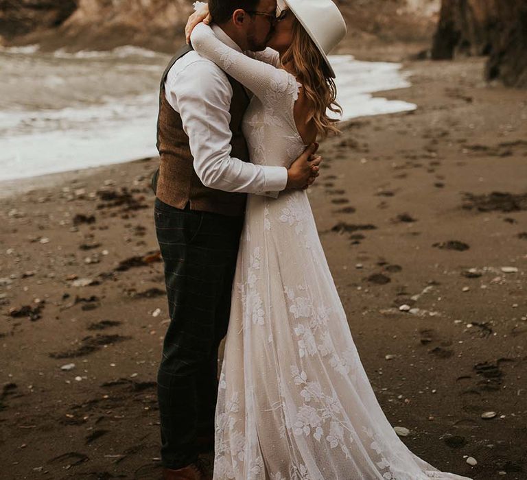 Intimate seaside elopement bride and groom portrait with bride in a long sleeve wedding dress and hat kissing her groom on the beach 