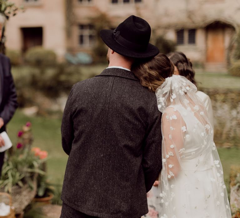 Bride in Charlie Brear wedding dress and daisy chapel length applique veil rests head on shoulder of groom in grey tweed suit and black fedora at garden party wedding at Hayne, Devon